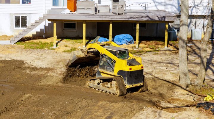 Backyard Grading and Landscaping Project with Skid Steer A yellow compact track loader moving soil in a backyard next to a house with a wooden deck, preparing the area for landscaping or drainage.