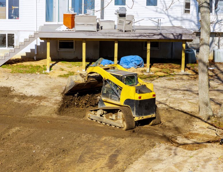 Backyard Grading and Landscaping Project with Skid Steer A yellow compact track loader moving soil in a backyard next to a house with a wooden deck, preparing the area for landscaping or drainage.