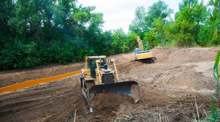 Slope Grading and Drainage Channel Construction A bulldozer grading a slope next to a muddy drainage ditch, with an excavator working in the background amidst dense green trees.