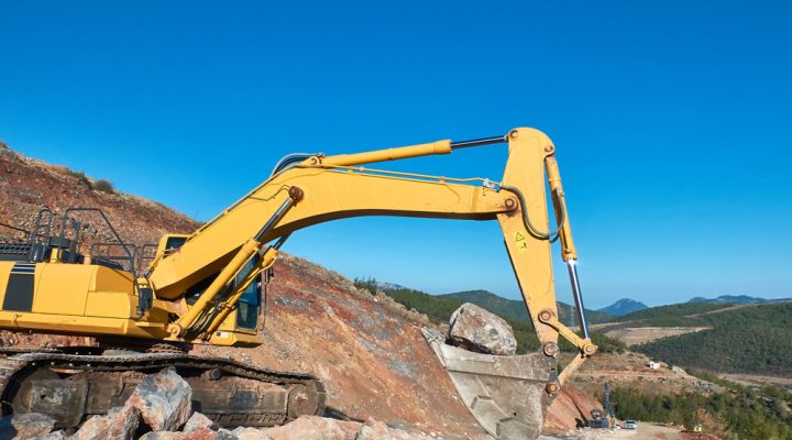 Heavy-Duty Rock Excavation and Demolition Services Large yellow excavator lifting a massive boulder at a rocky quarry site under a clear blue sky.