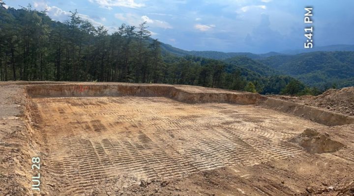 Large-Scale Mountain View Lot Grading for Custom Home Build A vast, meticulously graded building pad on a mountaintop with tire tracks across the soil, offering panoramic views of forested hills under a blue sky at 5:41 PM.