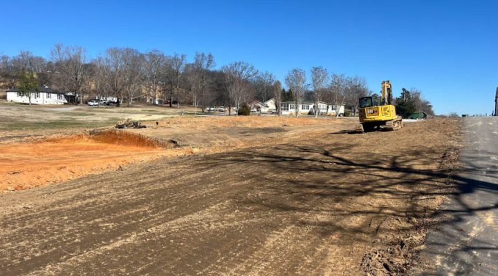 Roadside Grading and Soil Preparation for Development A yellow excavator parked on a graded roadside area next to a paved road, with a backdrop of suburban homes and bare trees under a clear blue sky.