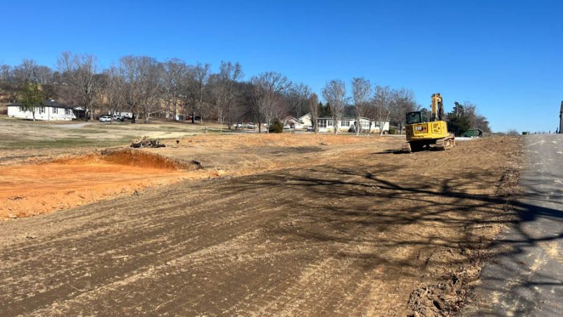 Roadside Grading and Soil Preparation for Development A yellow excavator parked on a graded roadside area next to a paved road, with a backdrop of suburban homes and bare trees under a clear blue sky.