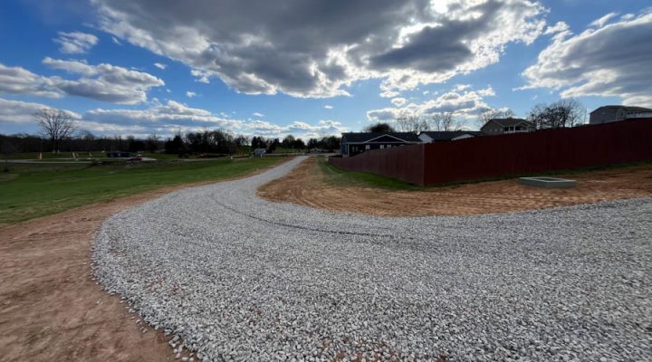 Newly Installed Gravel Driveway with Clean Edges and Professional Finish A winding gravel driveway bordered by freshly graded earth, leading towards a residential area under a dramatic sky with scattered clouds.