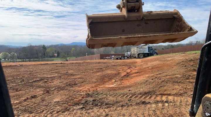 Heavy Equipment Hauling Soil from a Large Graded Site View from inside an excavator looking at its bucket raised high, with a dump truck parked on a large, graded red clay field ready to receive soil.
