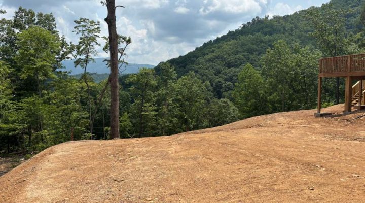 Cleared Hilltop Building Site with Panoramic Mountain Views A wide, cleared hilltop lot with a wooden deck structure on the right, offering expansive views of lush green mountains under a partly cloudy sky.