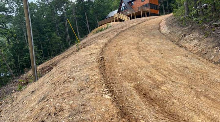 Completed Hillside Access Road Leading to a Modern Cabin A freshly graded dirt driveway winding up a steep hill to a modern wooden cabin, surrounded by dense forest under a cloudy sky.
