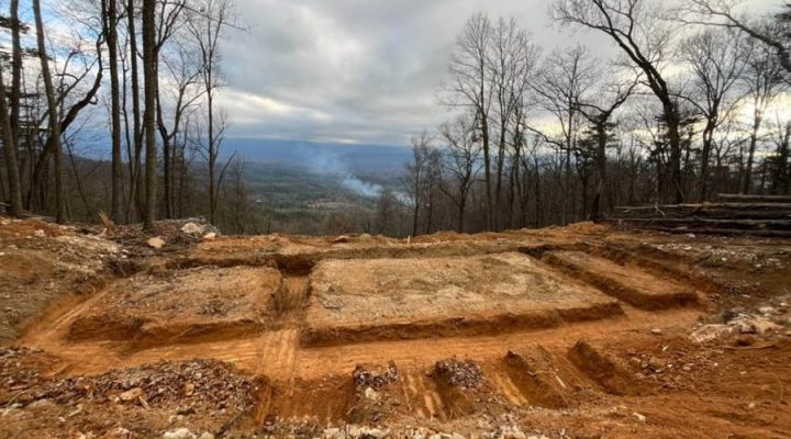 Foundation Trenching on a Mountain Top with Panoramic Valley Views Deep, red clay foundation trenches carved into a mountaintop site, surrounded by bare winter trees and offering a sweeping view of a distant valley under an overcast sky.