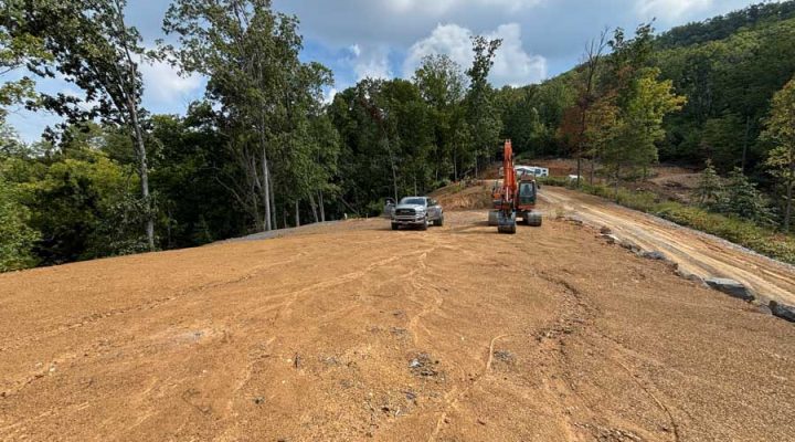 Large-Scale Land Clearing and Grading for Mountain Development A wide, graded lot with tire tracks, featuring an orange excavator and a pickup truck parked nearby, surrounded by dense trees and distant mountains under a cloudy sky.