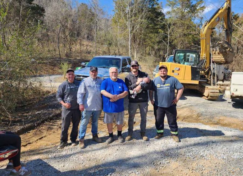 Rooney Excavating Team Photo on a Job Site Five members of the Rooney Excavating crew posing for a photo on a gravel site, with an excavator and truck in the background under a sunny blue sky.