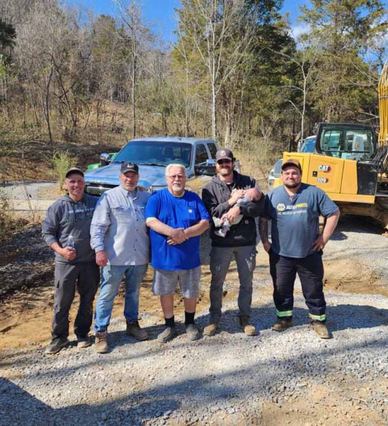 Rooney Excavating Team Photo on a Job Site Five members of the Rooney Excavating crew posing for a photo on a gravel site, with an excavator and truck in the background under a sunny blue sky.