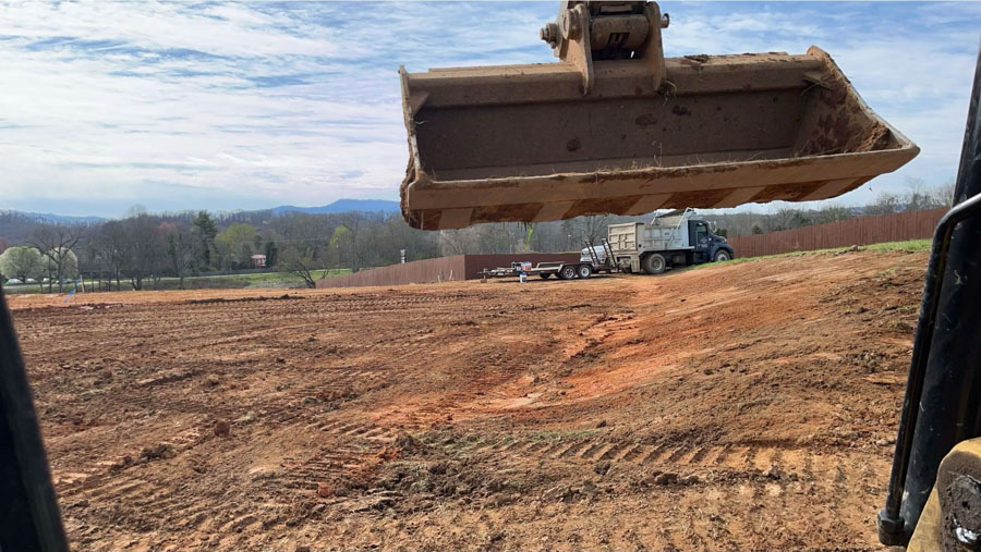 Excavator bucket raised over a large graded site, a truck and trailer in the background: site preparation by Rooney Excavating of Seymour, TN