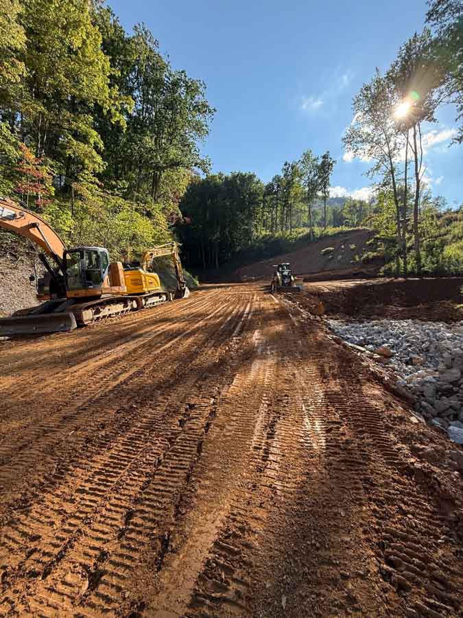 Excavator and a second machine grading a new road through a forested site, rock lining one edge: site preparation by Rooney Excavating of Seymour, TN