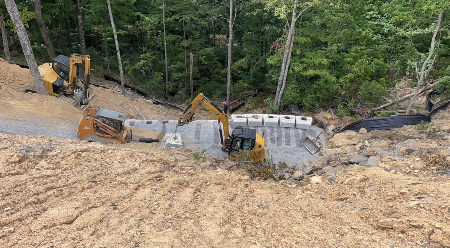 Excavator and skid steer working a steep wooded slope beside a stacked block wall: site preparation by Rooney Excavating of Seymour, TN